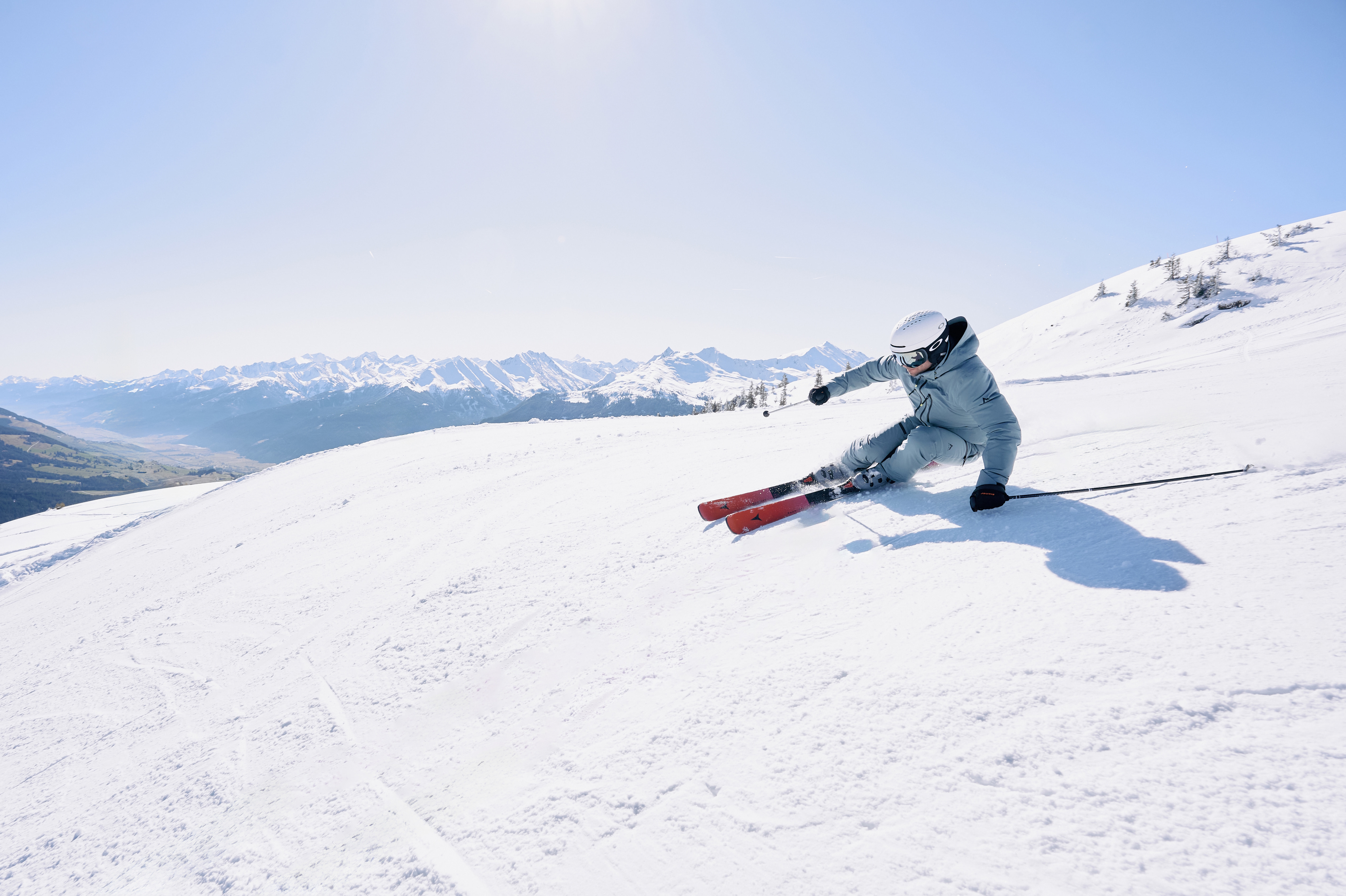 Eine Person fährt in dynamischer Kurve mit roten Skiern und grauer Skibekleidung eine schneebedeckte Piste hinunter, im Hintergrund erstreckt sich ein Gebirge unter blauem Himmel.