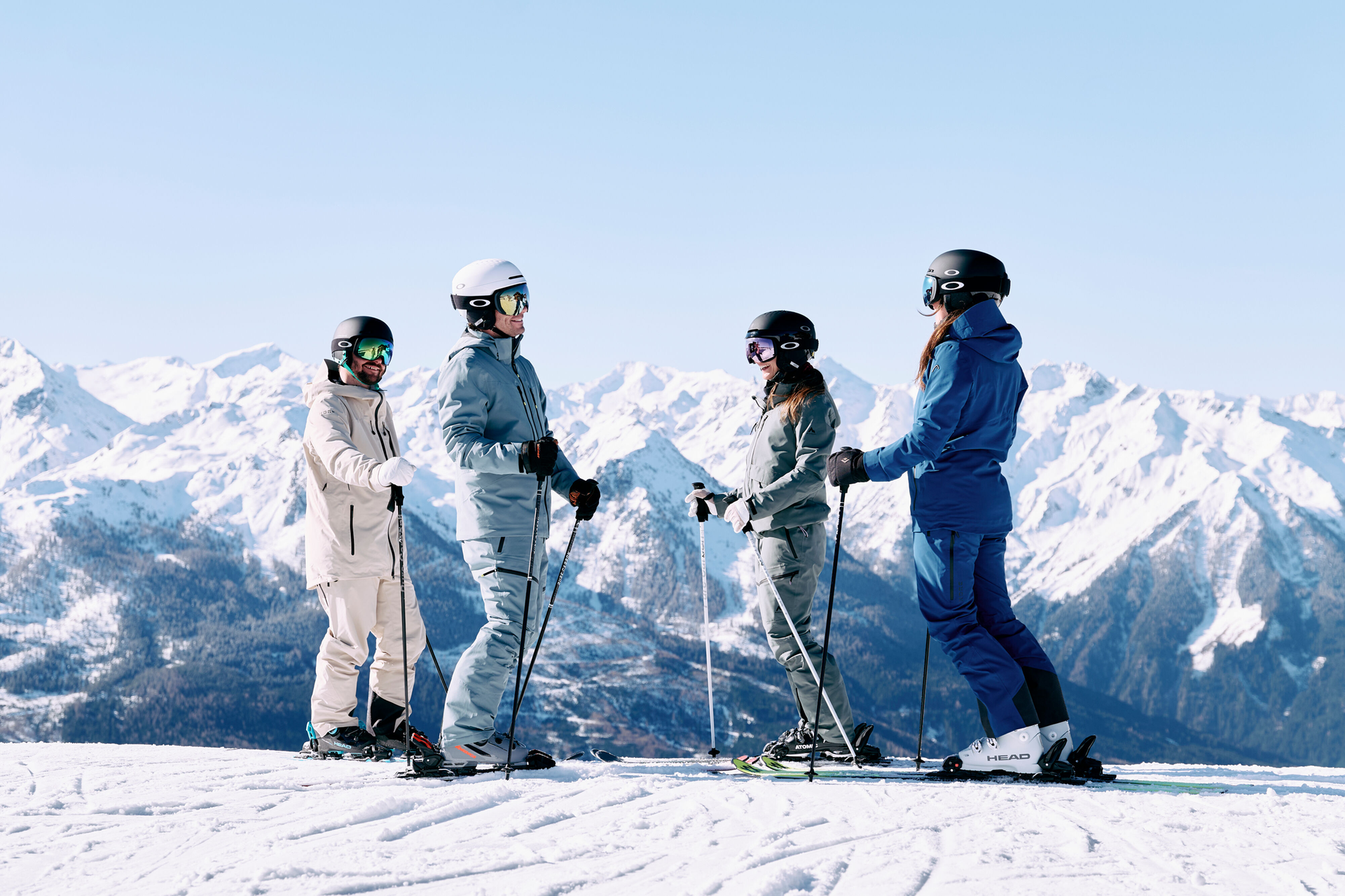 Vier Personen in Skiausrüstung stehen mit Skistöcken in den Händen auf einem verschneiten Berghang und blicken sich an. Im Hintergrund sind schneebedeckte Berge unter einem strahlend blauen Himmel zu sehen.