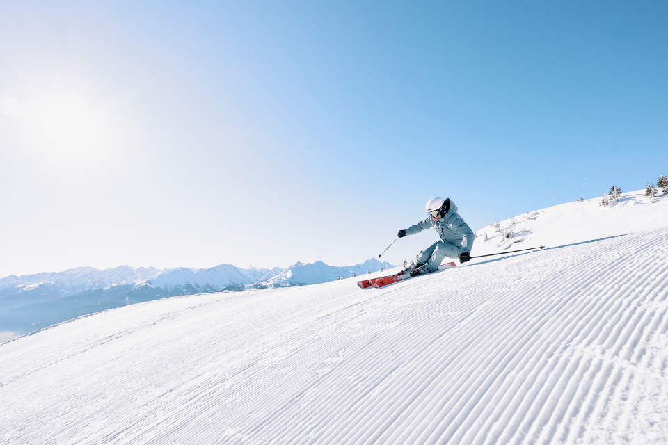 Ein Skifahrer in hellblauem Outfit und Helm fährt eine sonnenbeschienene, präparierte Skipiste hinunter, im Hintergrund sind Berge zu sehen, unter einem klaren blauen Himmel.