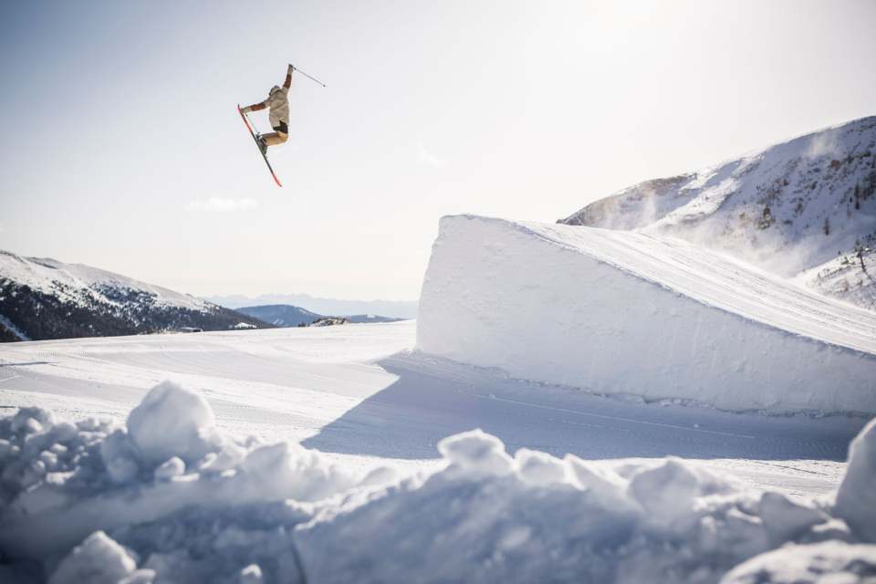 Ein Skifahrer führt in einer bergigen, verschneiten Landschaft einen Hochsprung von einer Schneerampe aus. Der Skifahrer schwebt mit gekreuzten Skiern vor einem klaren Himmel in der Luft.